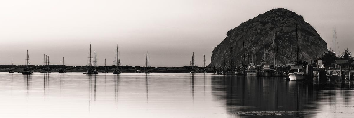 black and white modern wall art of Morro rock on the right side of the sand dune strand overlooking the morro bay with boats docked by the ramp